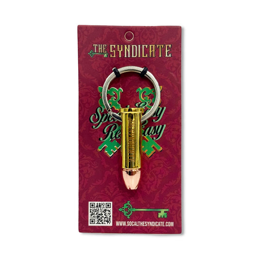 A gold-colored bullet-shaped key chain on a red background, with a small text that reads 'The Syndicate' and a website address.